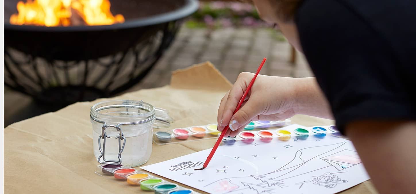A person is painting a picture with watercolors on a piece of paper by the fire pit