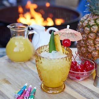 Tropical pina coladas with pineapple garnish and cherries on a table by a fire pit
