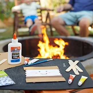 Craft supplies to make popsicle stick s'mores are sitting on a table next to a fire pit.