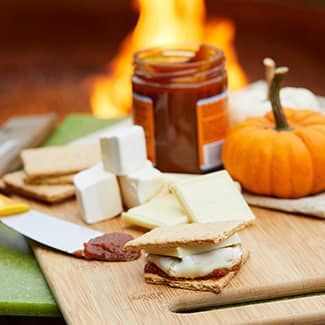 A wooden cutting board topped with s'mores ingredients for a pumpkin spice s'more, with fire pit