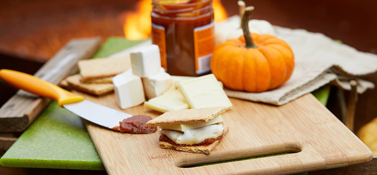 A wooden cutting board topped with s'mores , cheese , jam ingredients for a pumpkin spice s'more, and a pumpkin