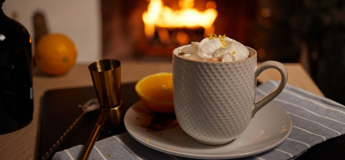 Spiked orange hot chocolate with whipped cream and orange zest, near an orange and a fireplace, fireside sip