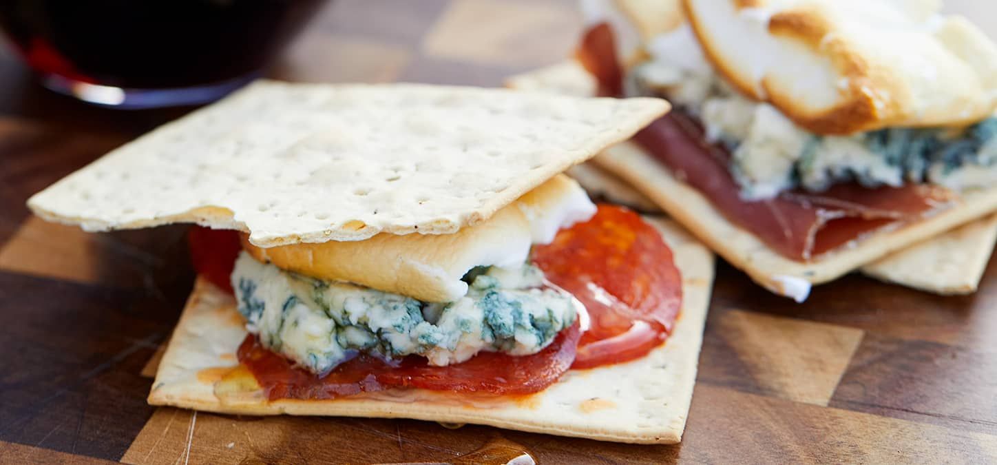 Savory smores with cheese and salami on a cracker with a glass of wine in the background.