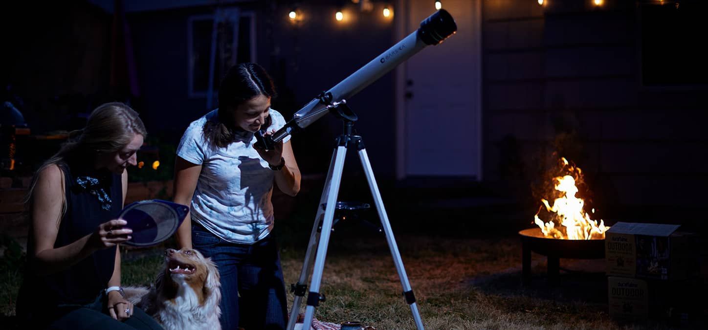 A woman is looking through a telescope at the night sky while a fire burns in fire pit in background
