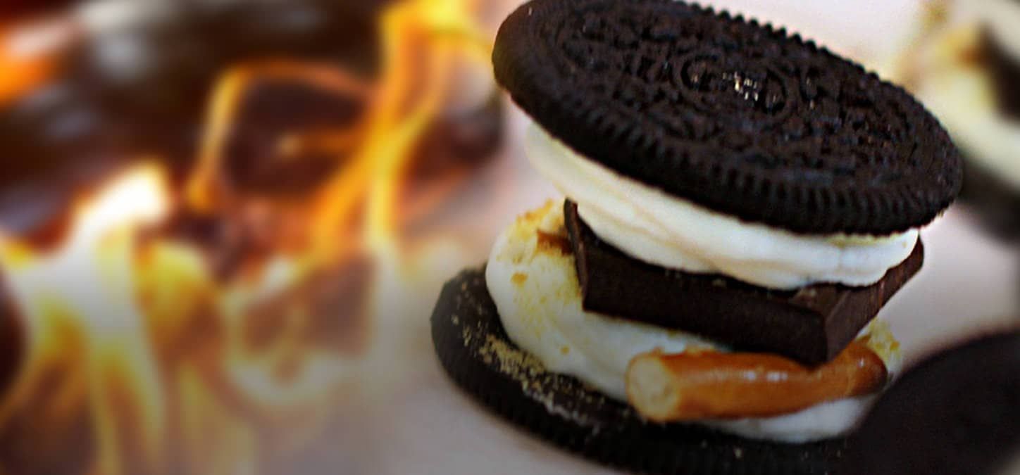 A close up of a sandwich cookie s'mores with chocolate marshmallow and pretzels with a fire in the background.