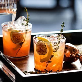 Two spiced fig bourbon sour cocktails with lemon and herbs on a tray, with a copper shaker beside them.