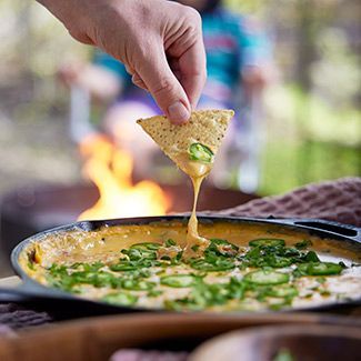 Hand dipping a tortilla chip into a skillet of cheesy dip with jalapeños, by a campfire.