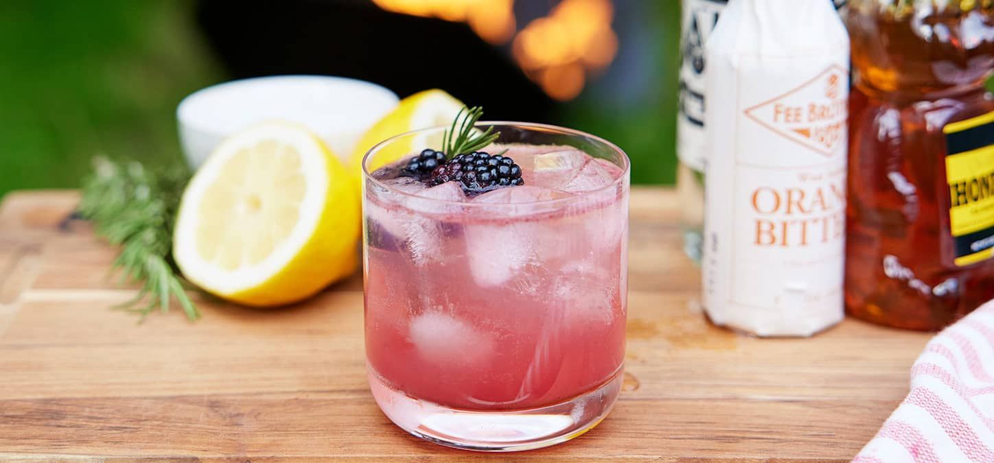 A close up of a spooky blackberry smash cocktail in a glass on a table in front of a fire pit