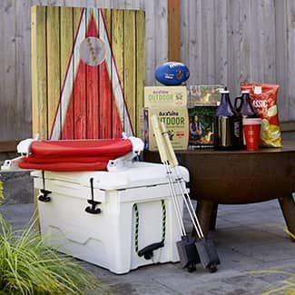 A cooler with red seat, cornhole game, food, beer, and fire pit.