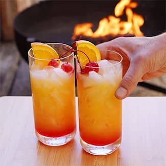 Two tequila sunrise cocktails with ice and lemon slices, with a fire in the background
