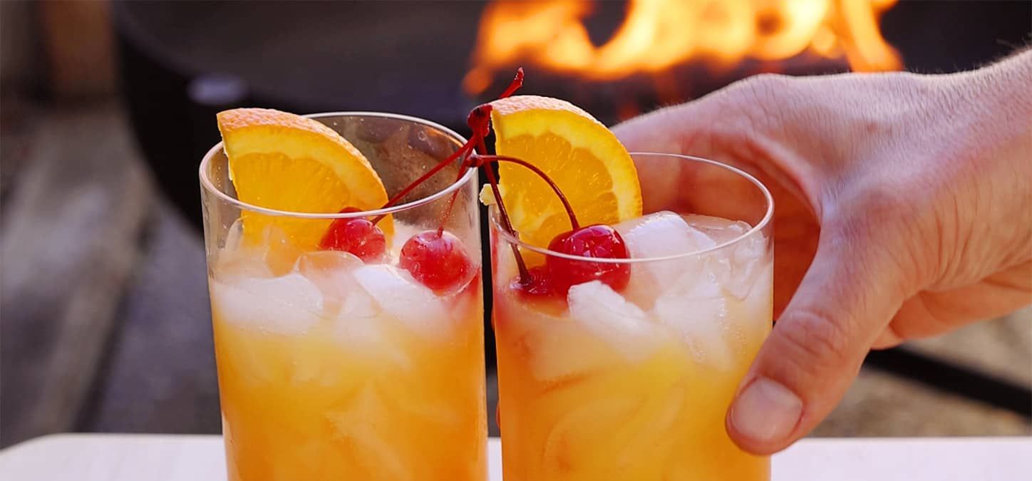  two glasses of tequila sunrise garnished with cherries and oranges in front of a fire pit