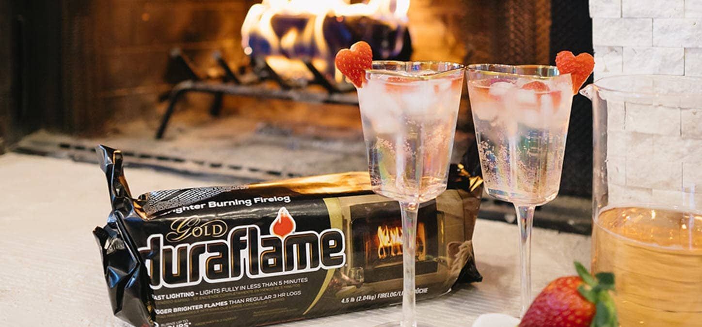 two Valentine's day sangrias garnished with heart-shaped strawberries is next to a package of durafllame Gold firelog with a fire in the fireplace.