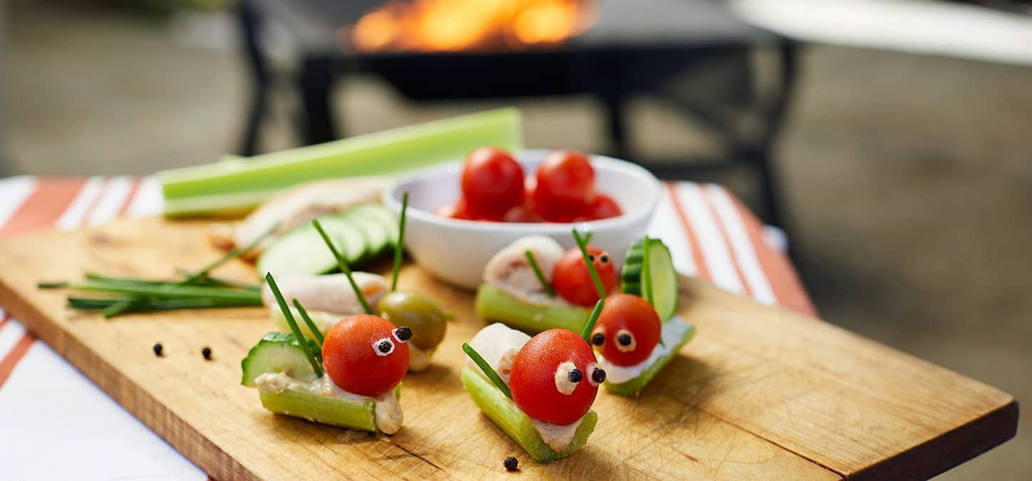 A wooden cutting board topped with tomatoes , cucumbers , olives and cheese shaped into grasshoppers