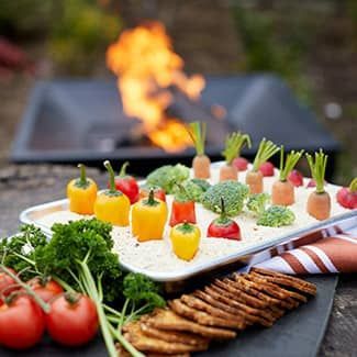 A tray of vegetables and crackers on a table with a fire pit in the background.