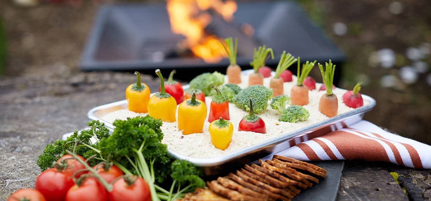 A tray of vegetables and crackers on a table with a fire pit in the background.