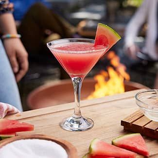 Watermelon cocktail in a martini glass beside a fire pit with watermelon slices on a table