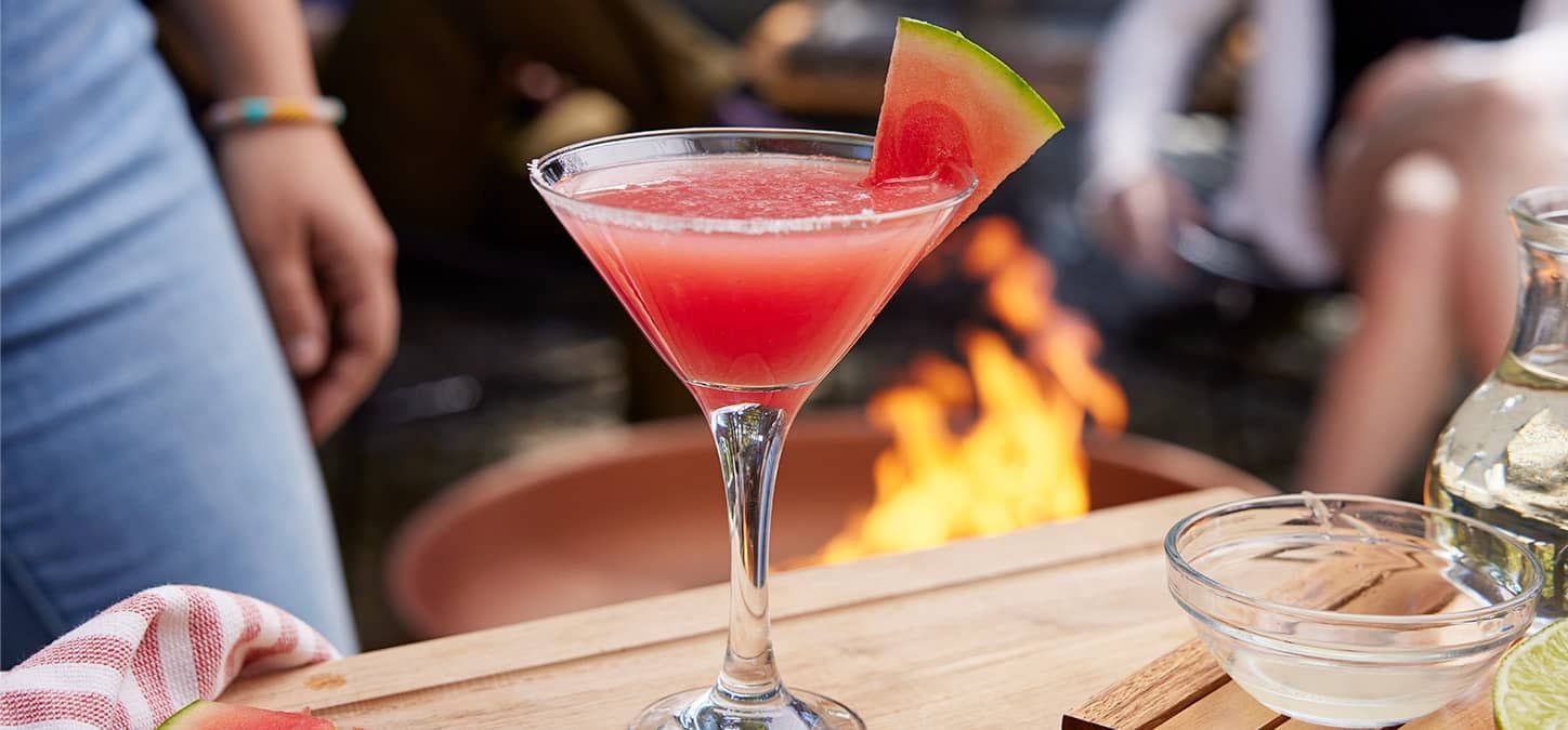 A watermelon martini with a slice of watermelon garnish with a fire pit in the background