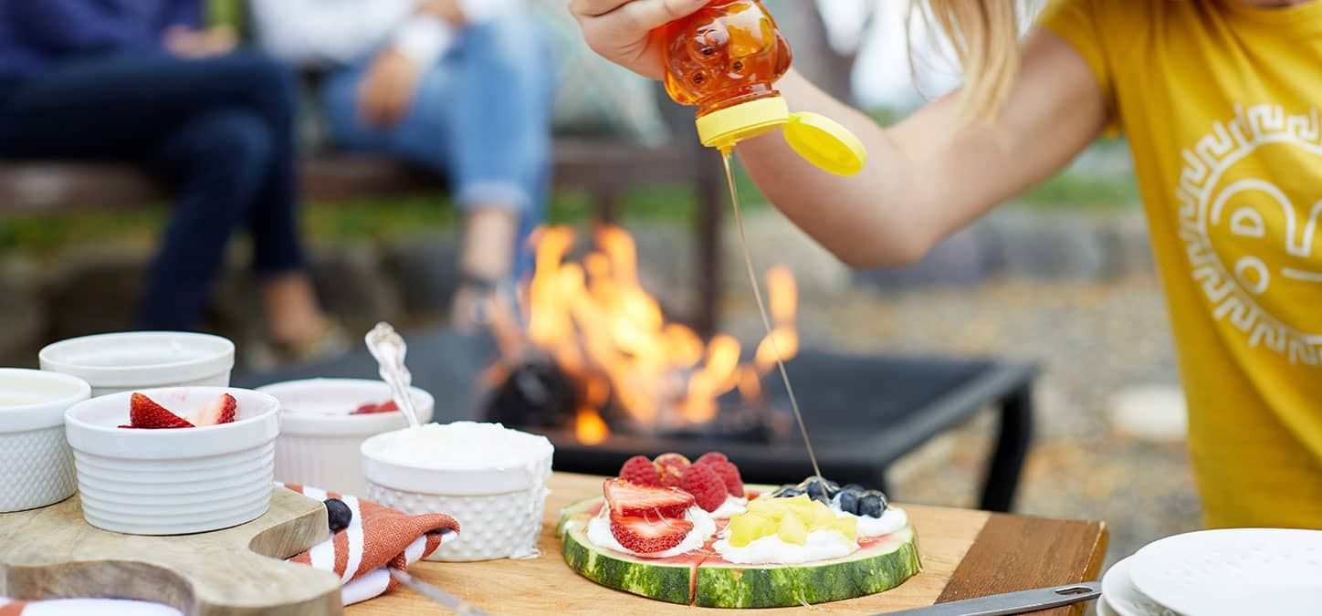 A person is pouring honey on a watermelon slice with a fire pit in the background
