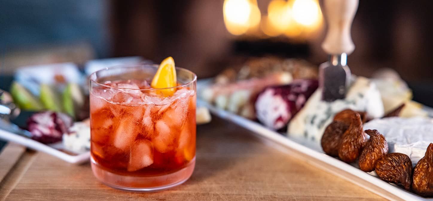A close up of a negroni with ice and a lemon wedge on a table next to a plate of appetizers