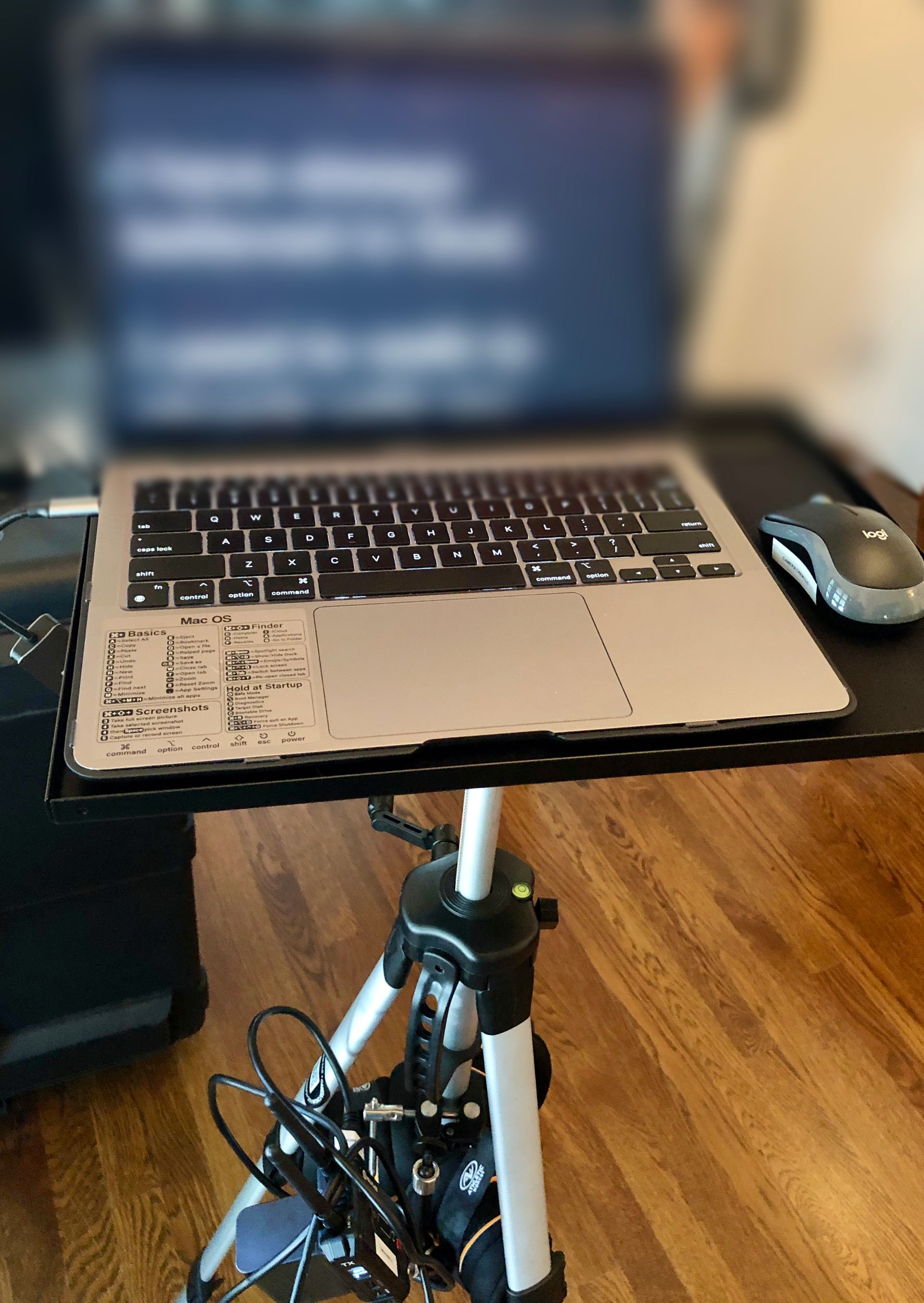 A laptop sits on a tripod stand with a computer mouse beside it on a desk, set against a wooden floor background.