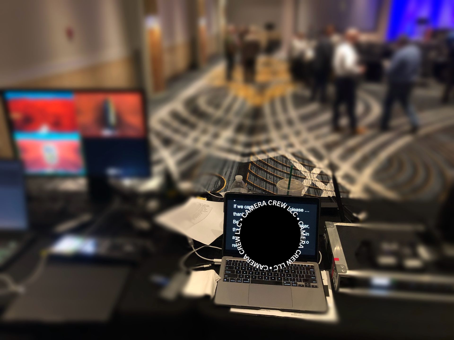 A laptop and equipment on a table, with a blurred conference room and attendees in the background.