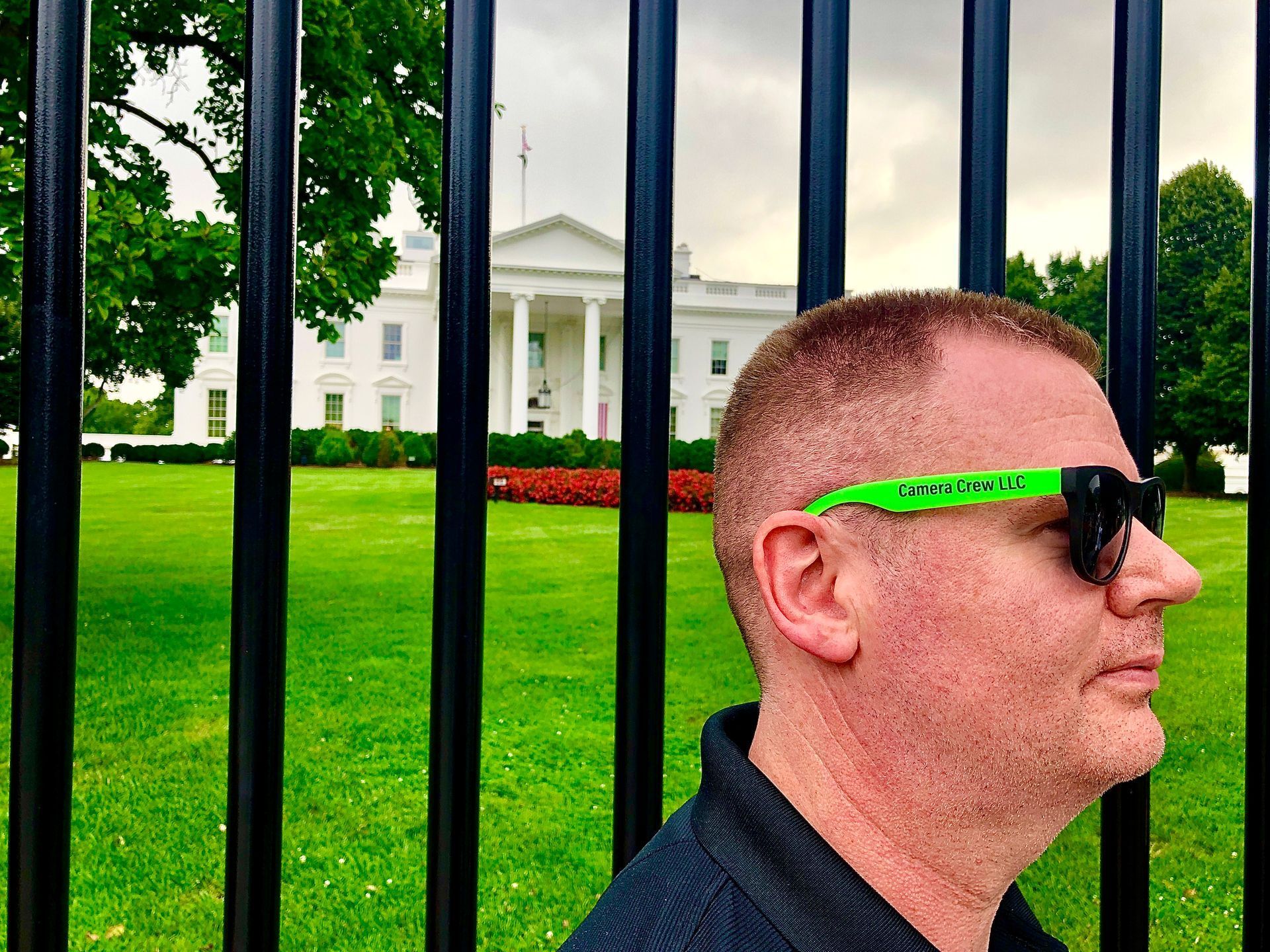 A person wearing bright green sunglasses stands in profile before a black fence with the White House in the background.