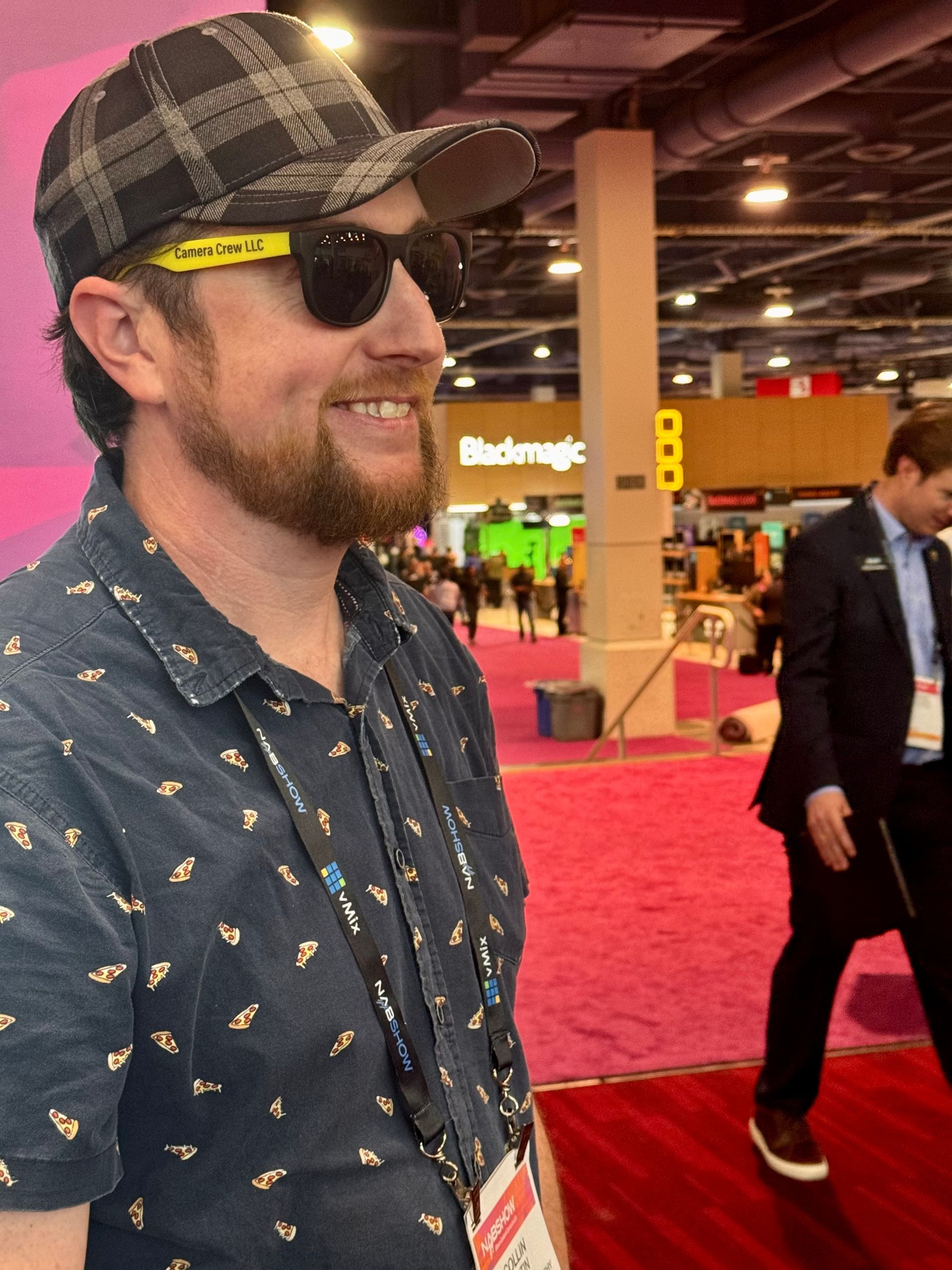 A smiling person in a plaid cap and patterned shirt walks past a Blackmagic Design booth in a brightly lit convention hall.
