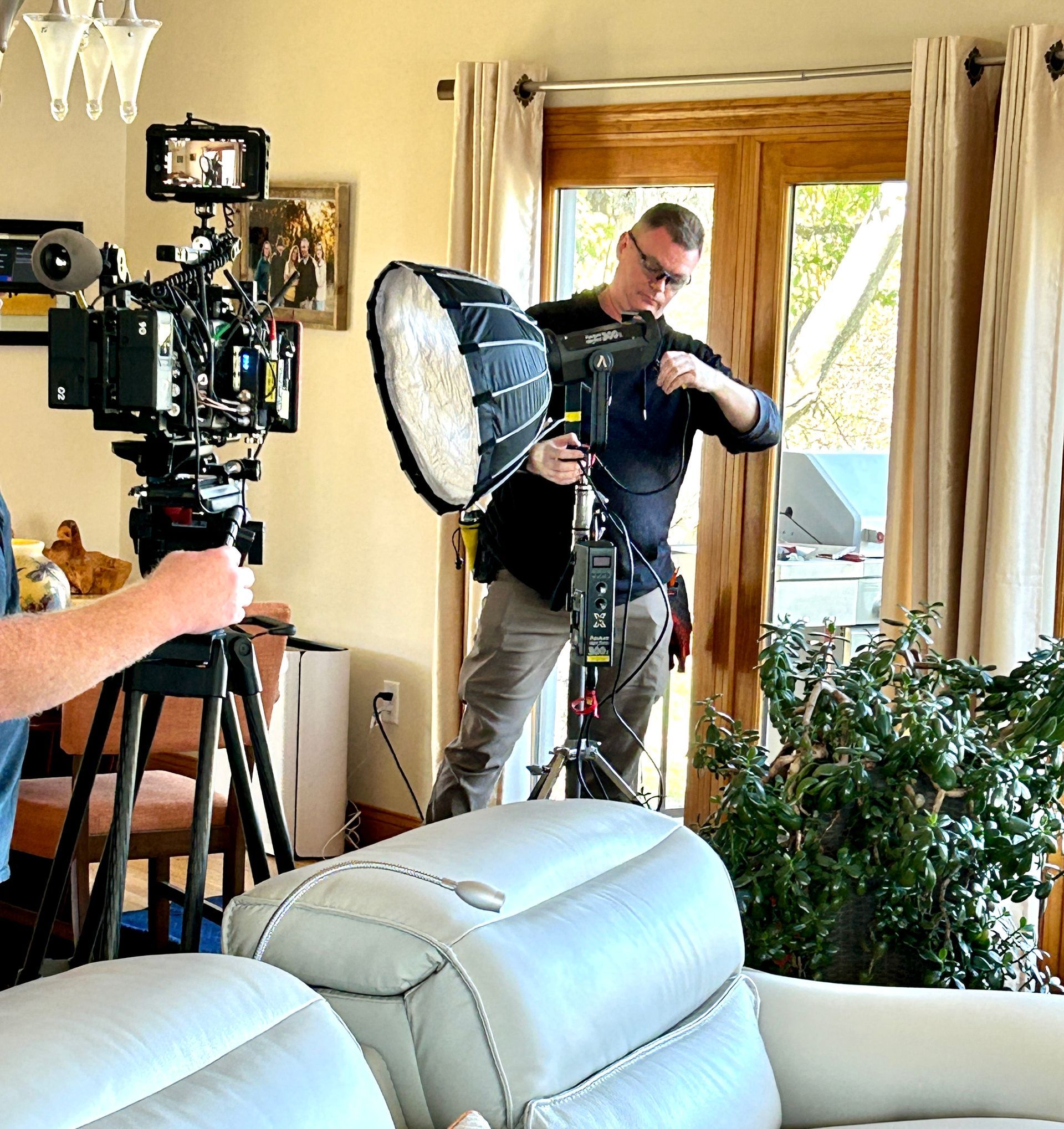 A person adjusts a studio light with a softbox near a window while a camera on a tripod films the scene in a living room.