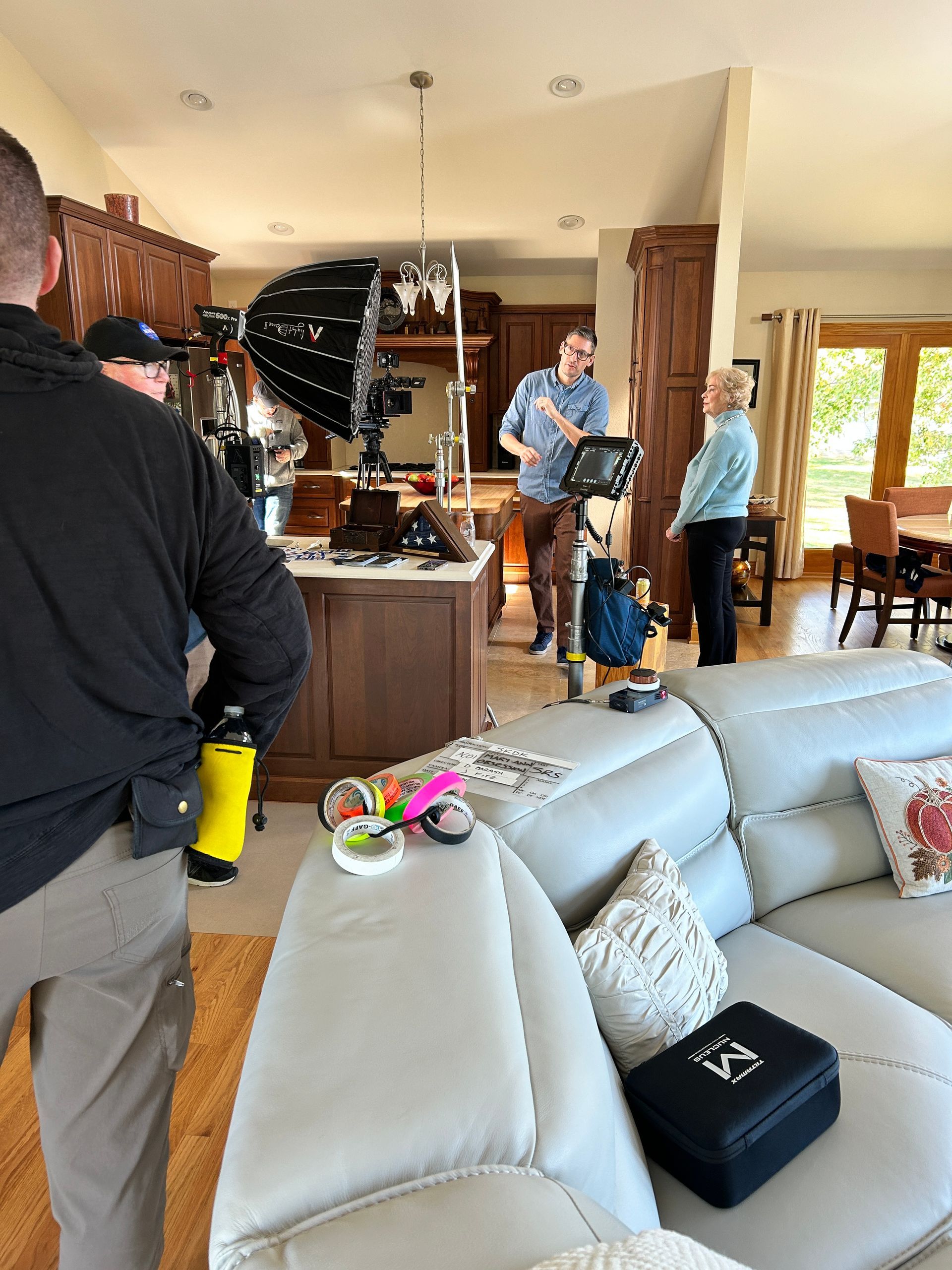 A film crew setting up lighting equipment in a kitchen and living area, with three people standing and conversing.
