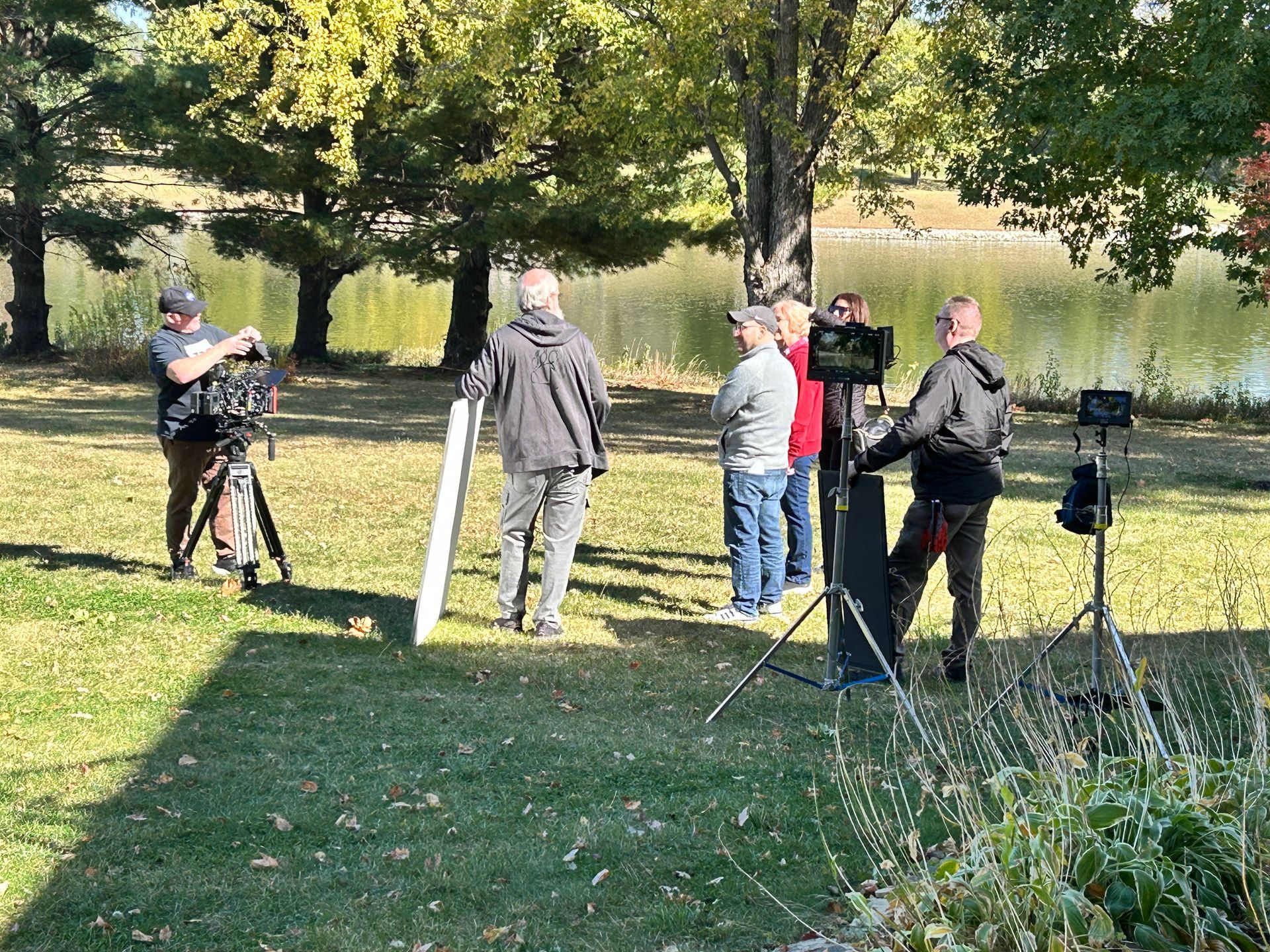 A film crew captures a scene outdoors by a lake, with camera operators, a reflector, and individuals standing on grass.