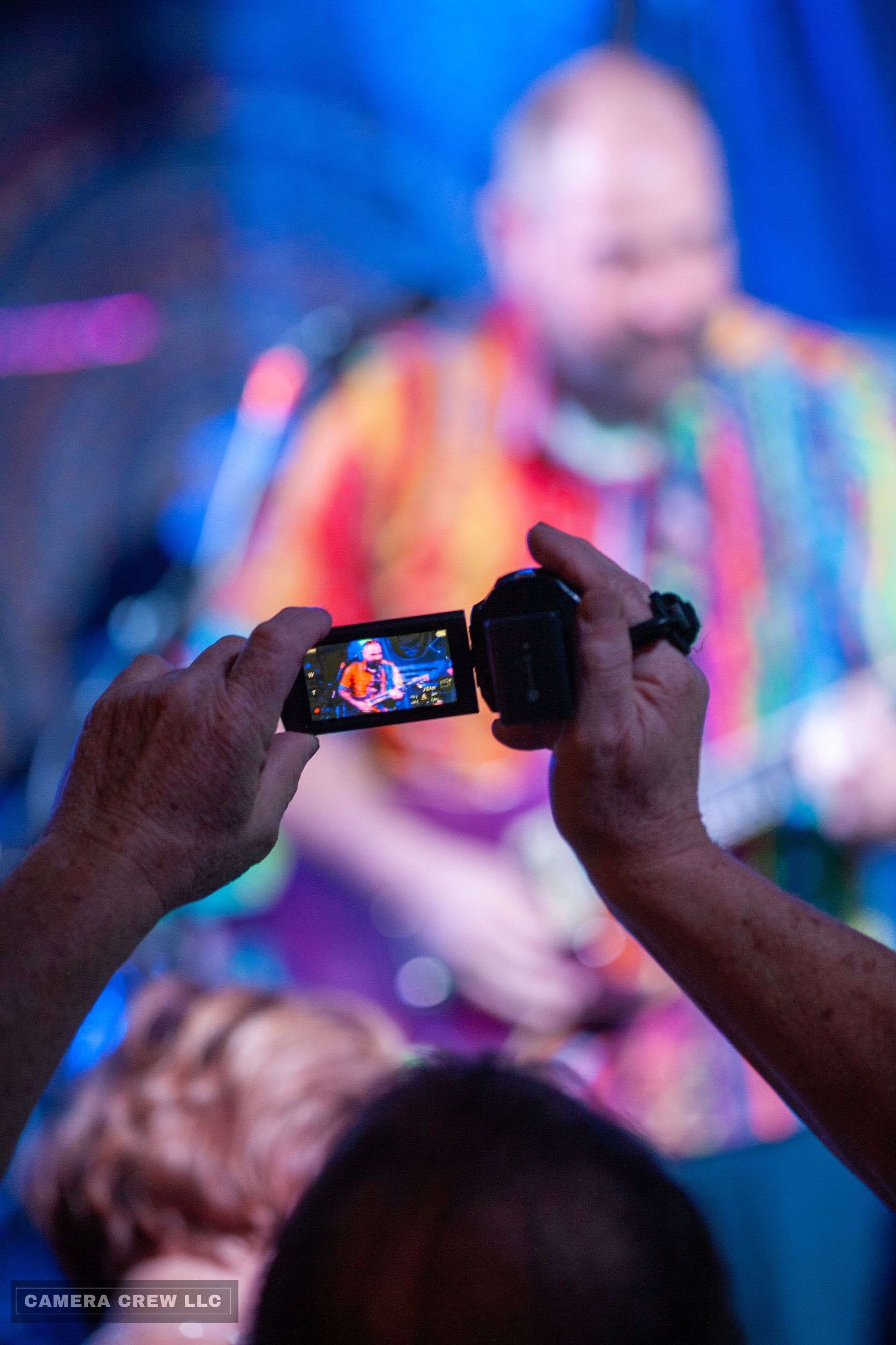 A pair of hands holds a camcorder to record a person wearing a vibrant, multicolored shirt on a stage with blue lighting.