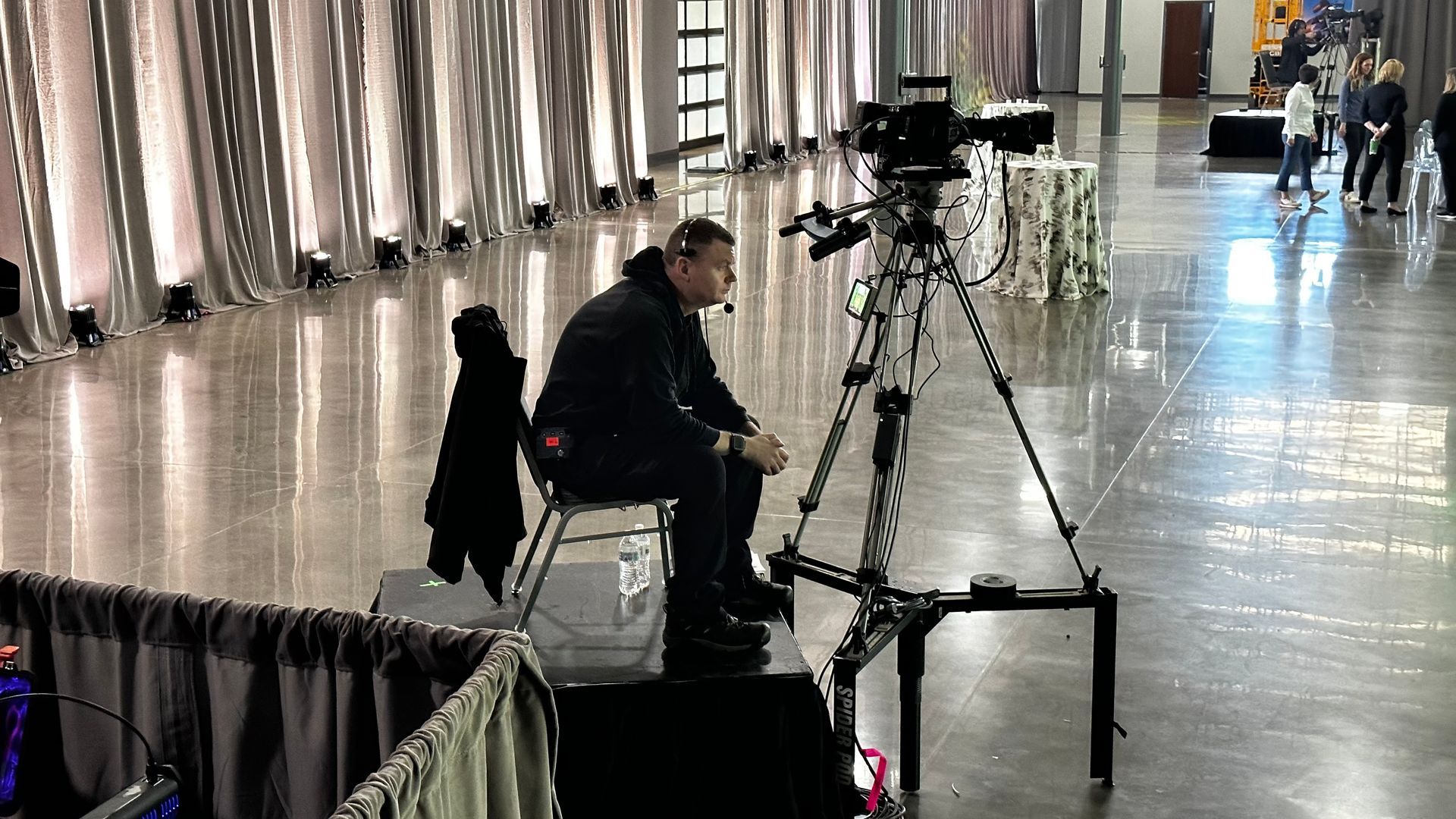A videographer sits on a platform behind a professional camera on a tripod in a large, modern event space.