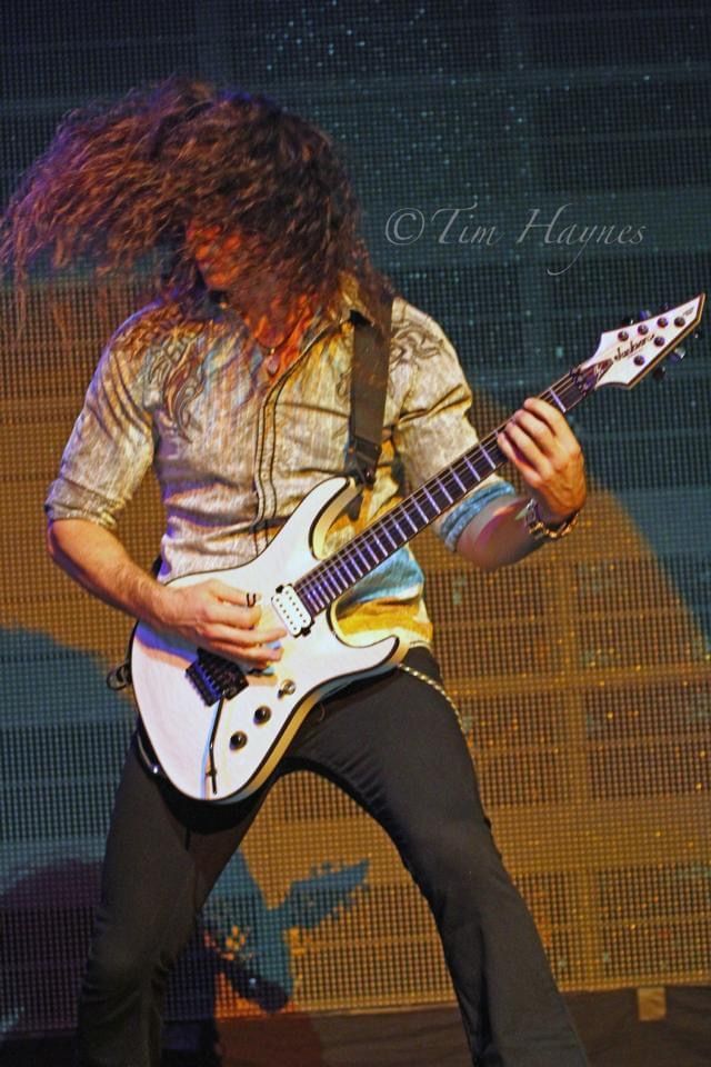 A long-haired guitarist on stage, headbanging while playing a white electric guitar in front of a golden backdrop.