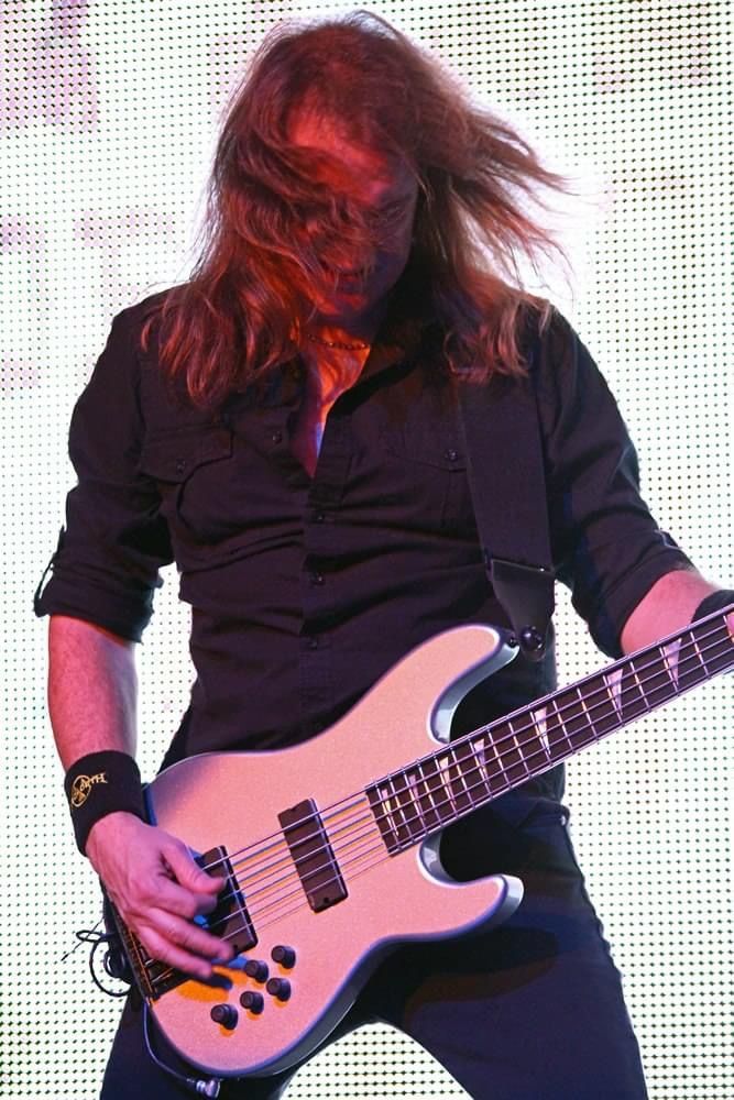 A guitarist with long hair playing a light-colored electric guitar on a brightly lit stage with a grid-pattern backdrop.