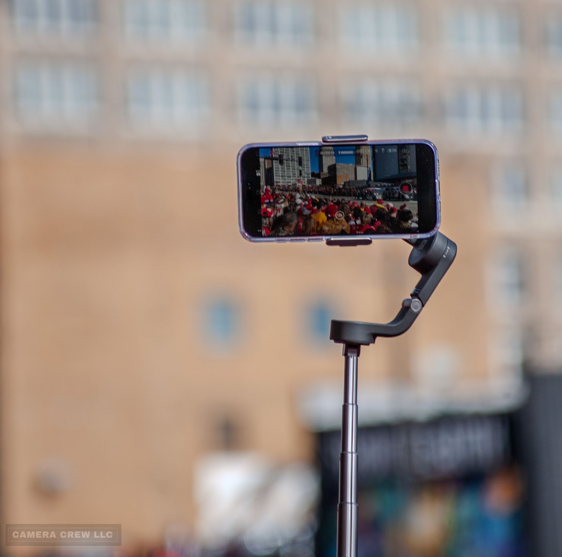 A smartphone mounted on a gimbal stabilizer recording a blurred outdoor crowd against a blurry building background.