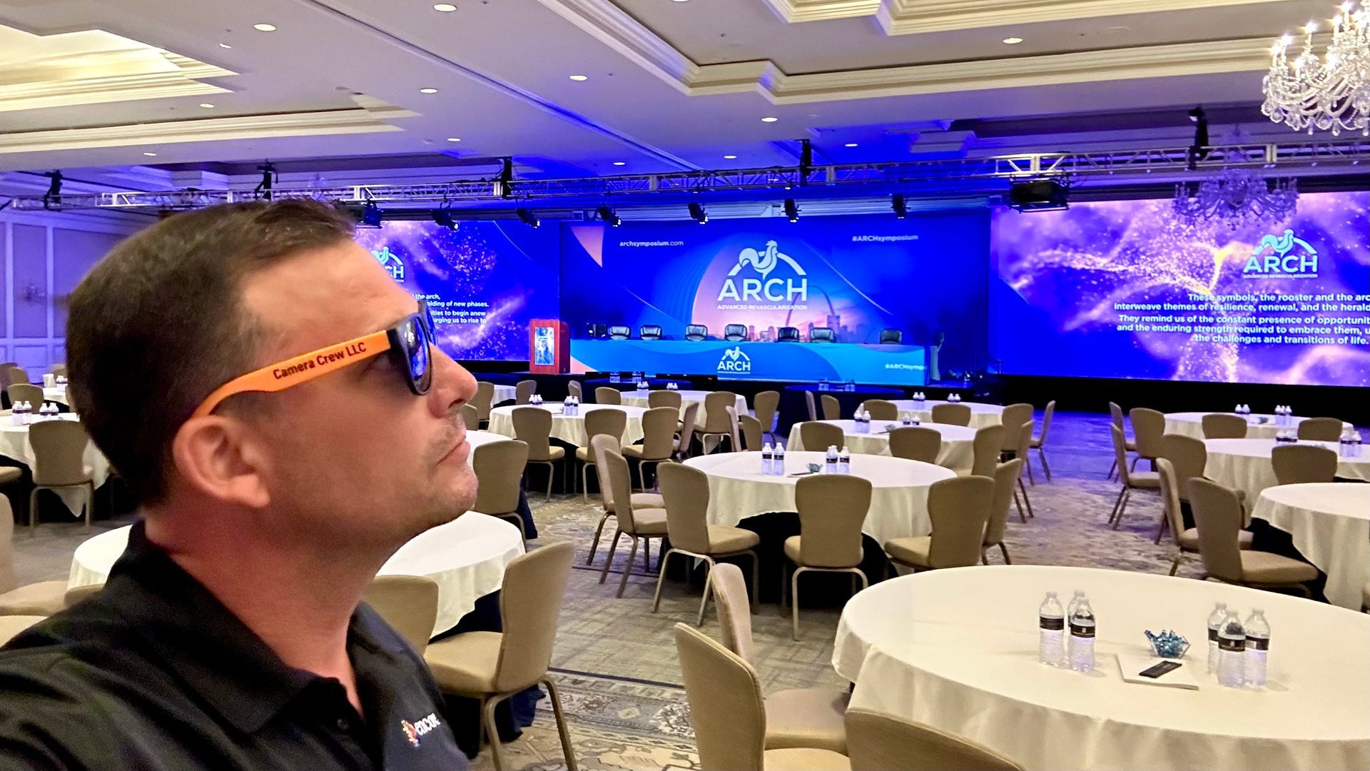 A person wearing orange sunglasses in a large, brightly lit ballroom with round tables set for a conference.