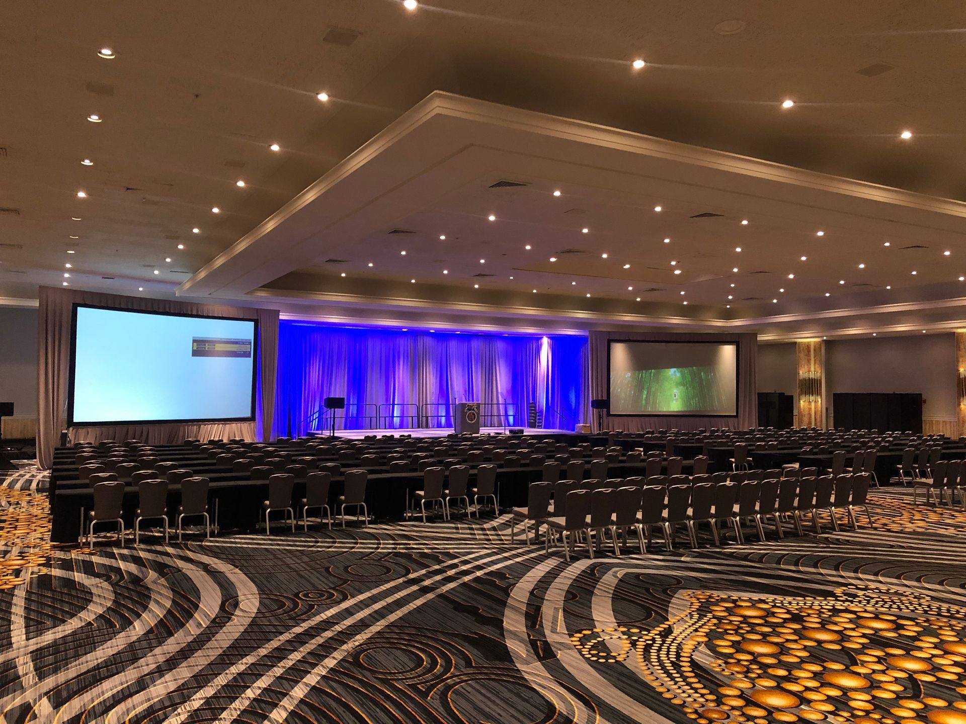 An empty hotel ballroom set for a presentation with two large screens, stage lighting, and rows of chairs and tables.