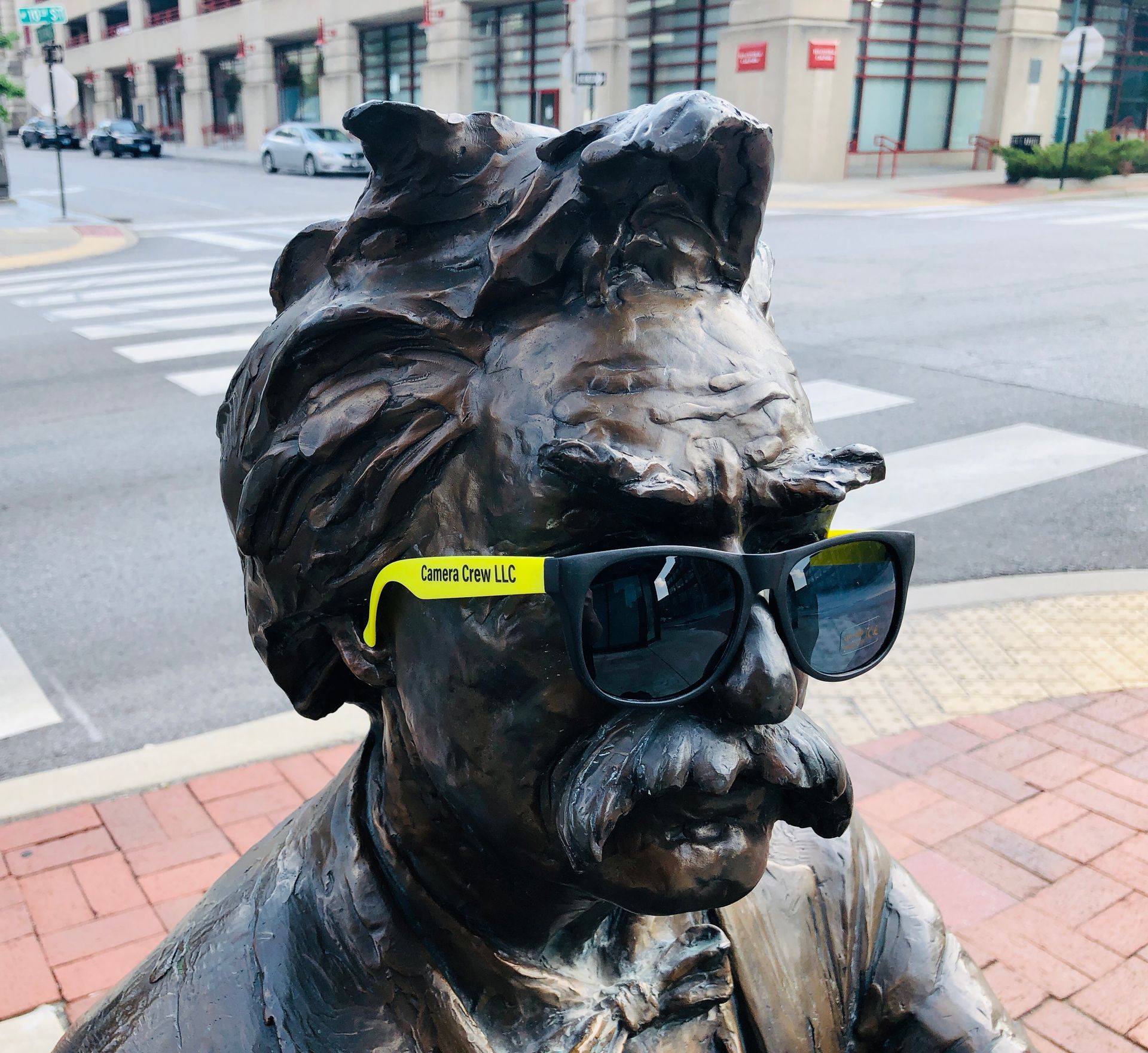 A bronze statue of Mark Twain wearing bright yellow-templed sunglasses on a sidewalk near an intersection.
