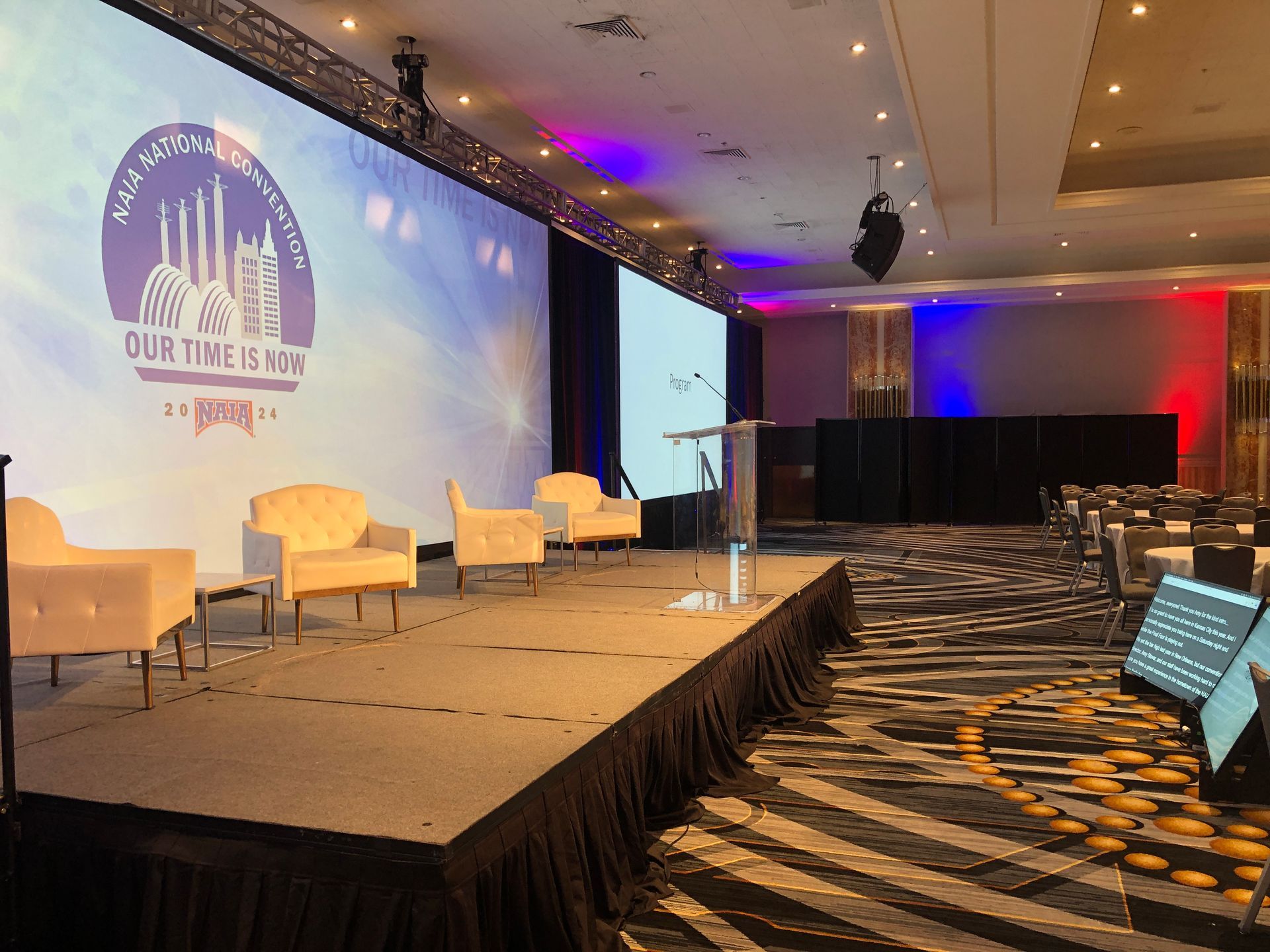 An empty stage with chairs and a podium in a banquet hall, featuring a large screen with a logo and decorative lighting.