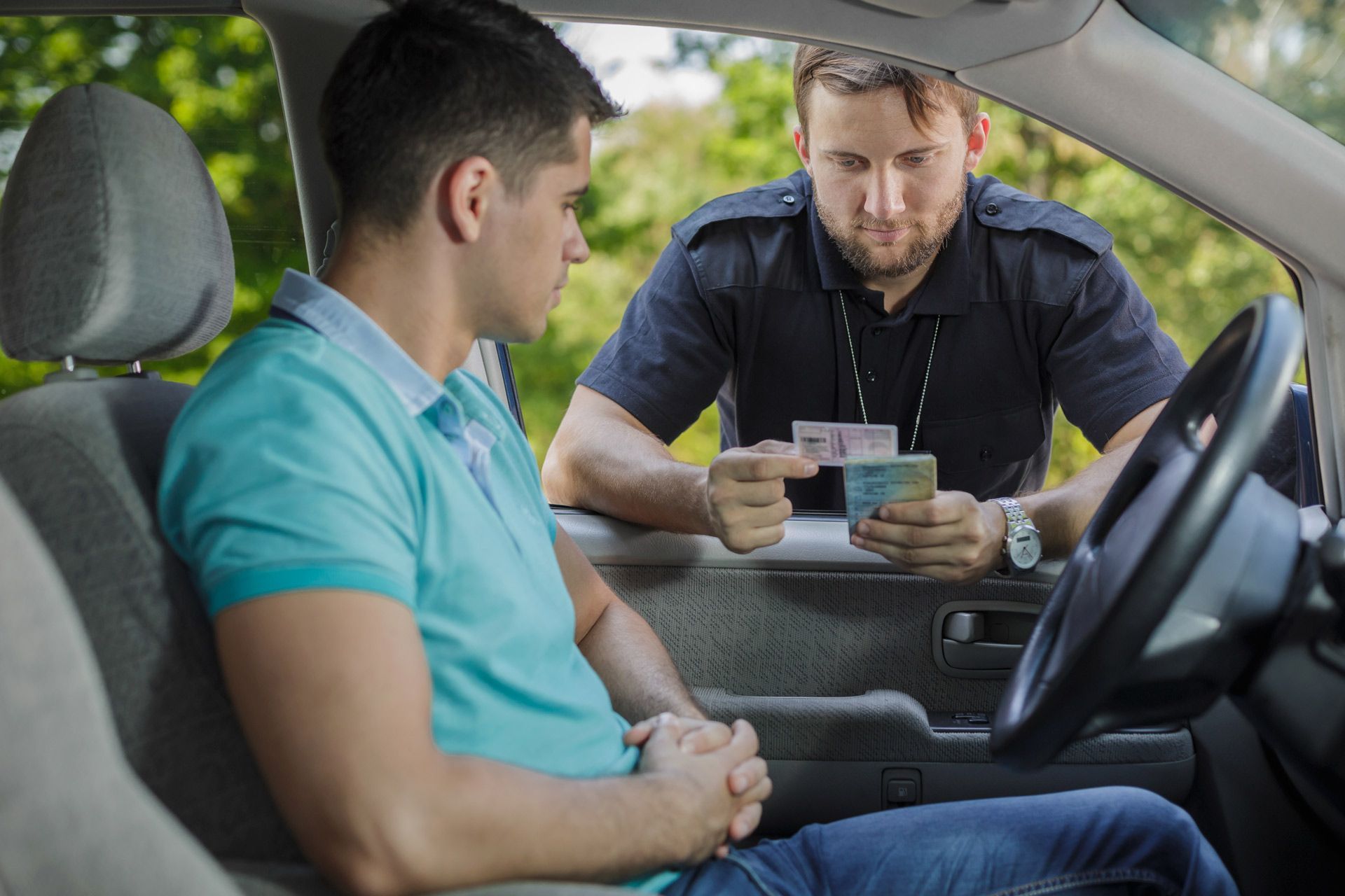 Police officer examining a driver's license; driver in car looks concerned.