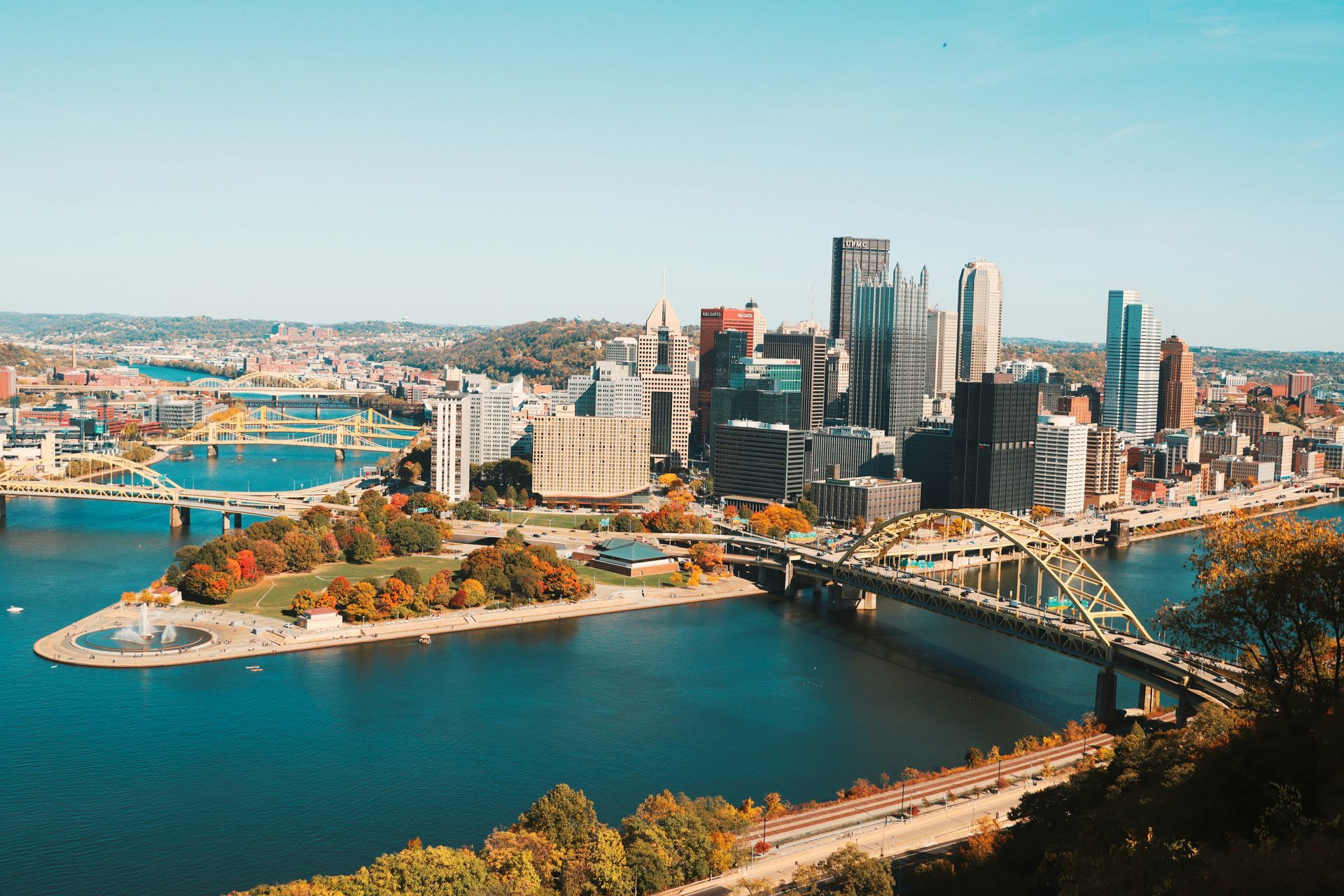 Pittsburgh skyline with bridges over rivers, buildings, and a park, under a blue sky.