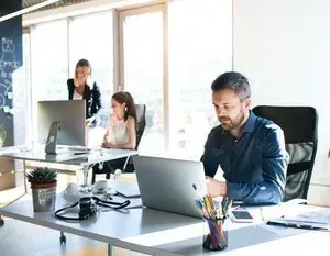People working at desks in a bright office; man typing on a laptop, others at computers.