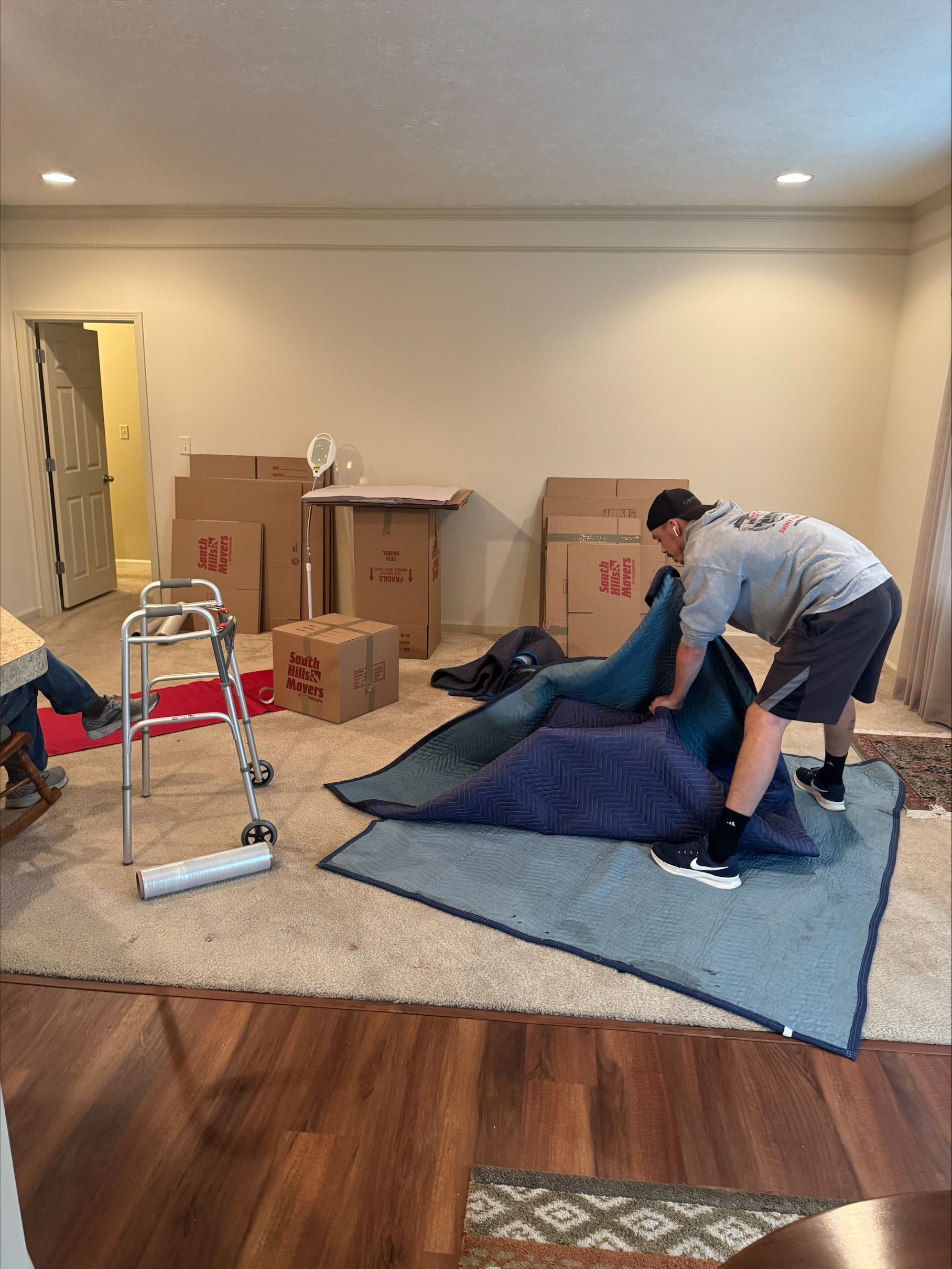 Man lifts a wrapped blue cushion in a room with moving boxes, a walker, and hardwood floors.
