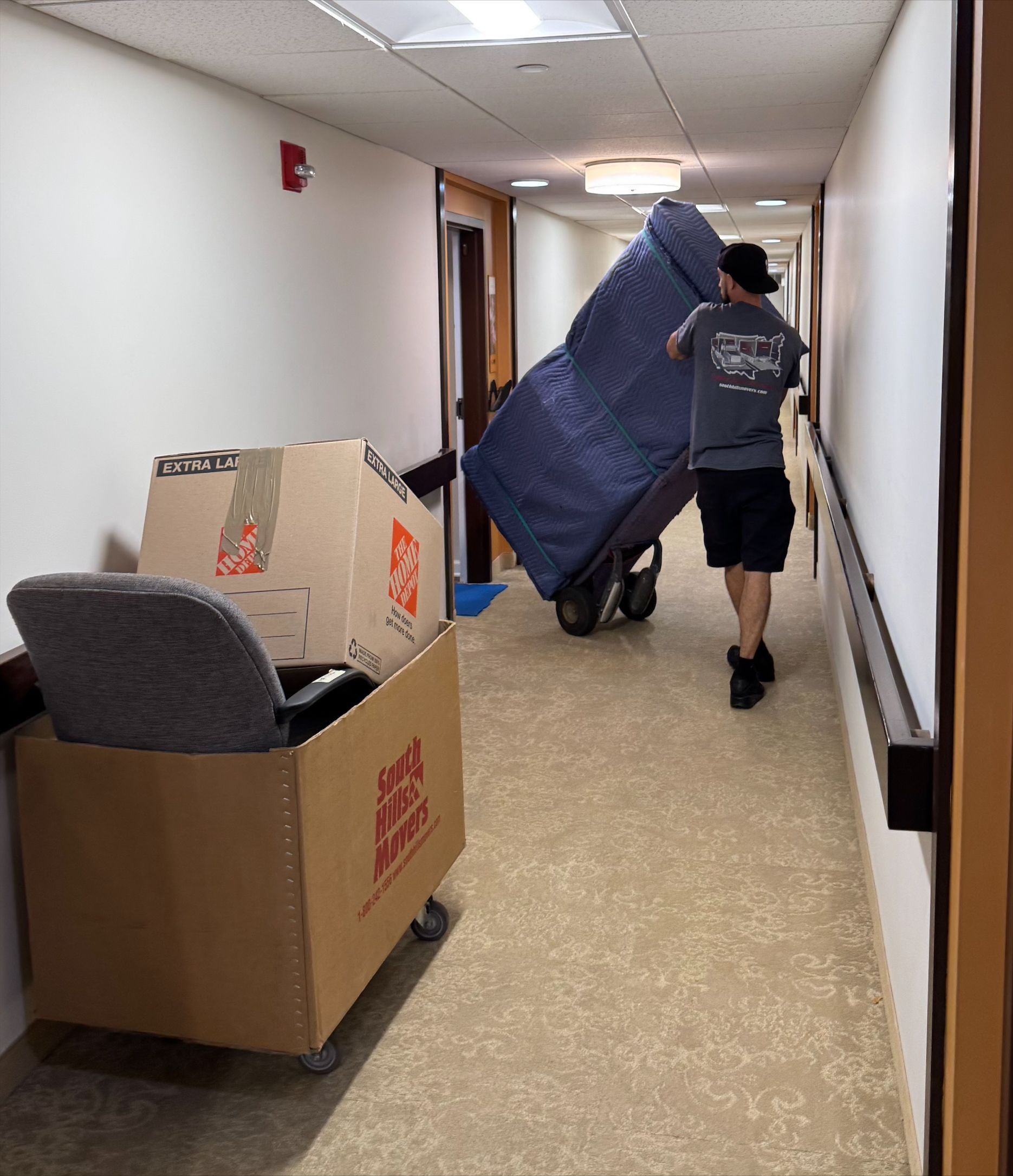 Man moving furniture on a dolly down a hallway. Boxes and a chair are also in the hallway.