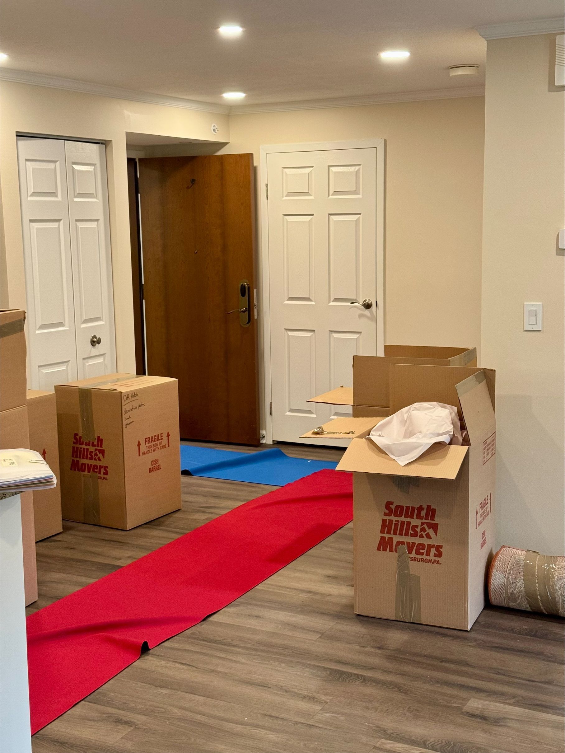 Boxes and red carpet inside, next to a door, in a room with hardwood floors.