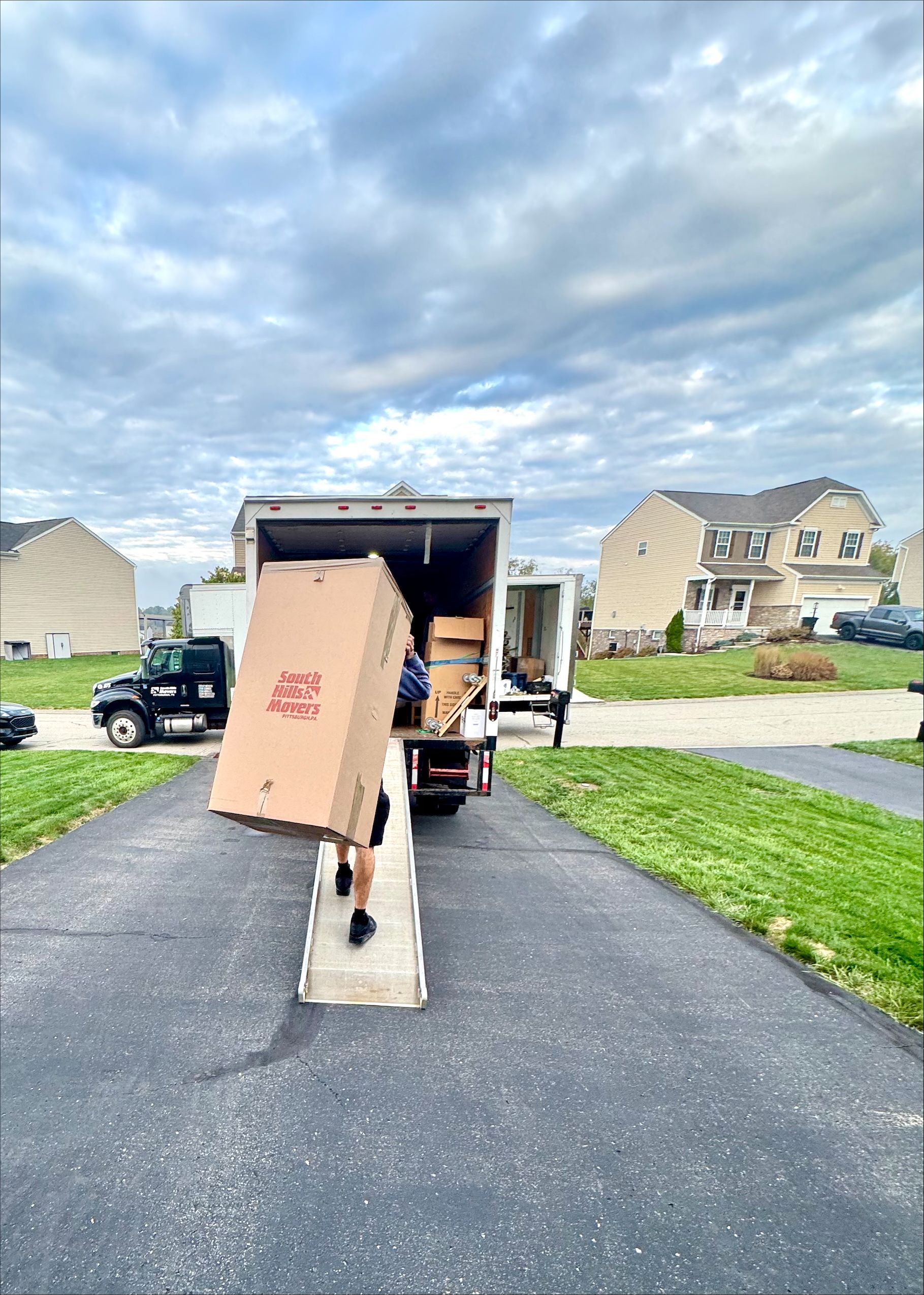 Person carrying a large cardboard box from a moving truck onto a driveway, residential neighborhood.