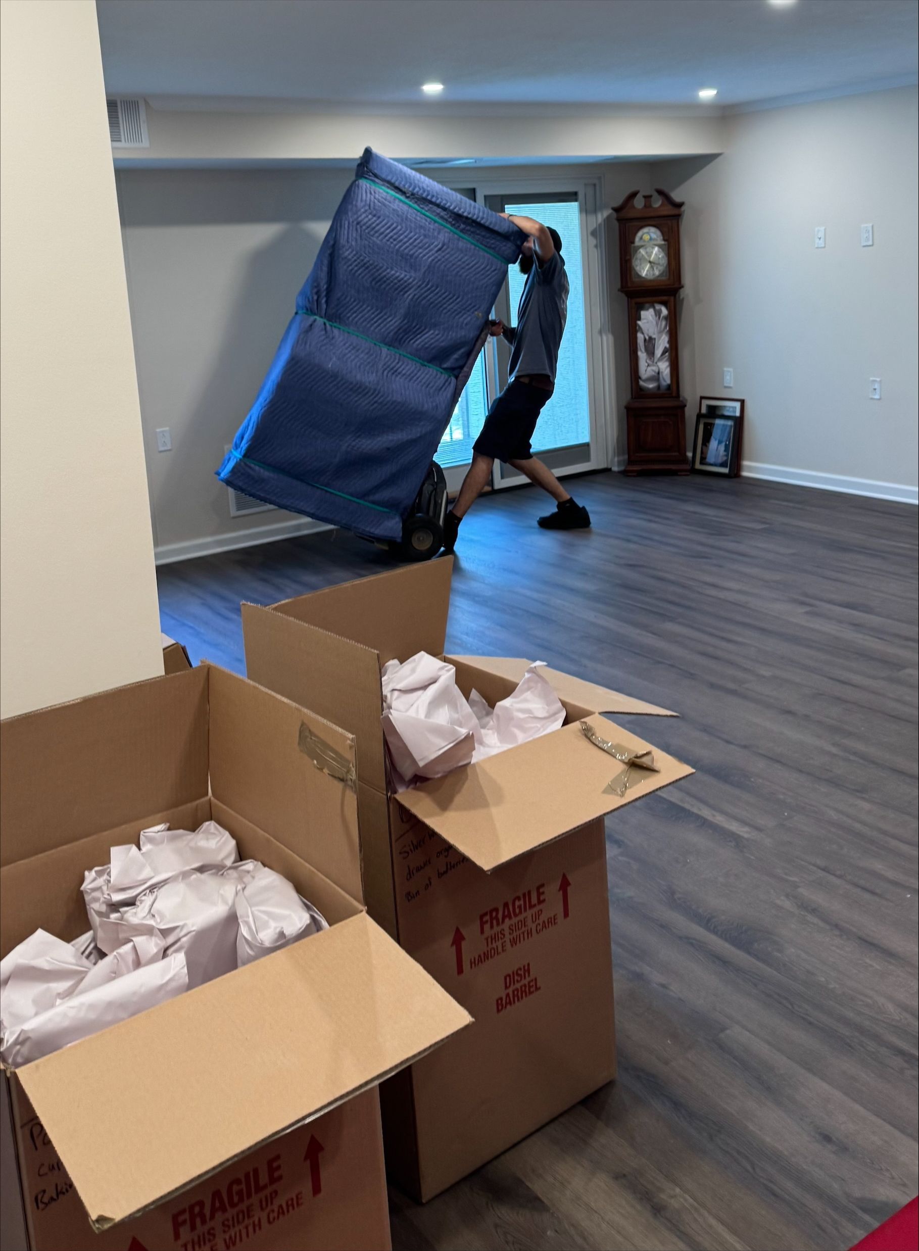 A person carrying a large, blue-covered object in a room with boxes and a grandfather clock.