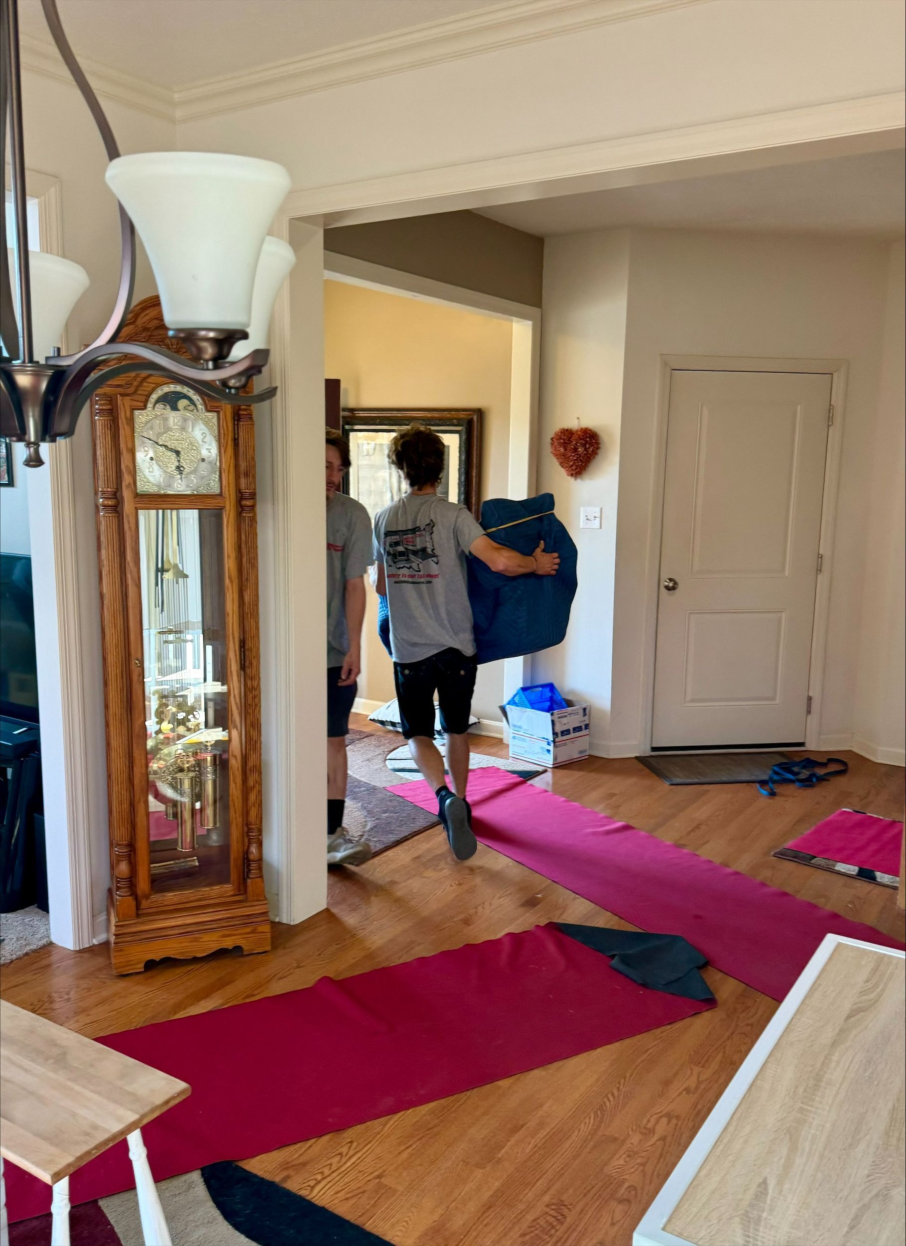 Two people carrying a blue item through a doorway into a room with hardwood floors and a red runner.