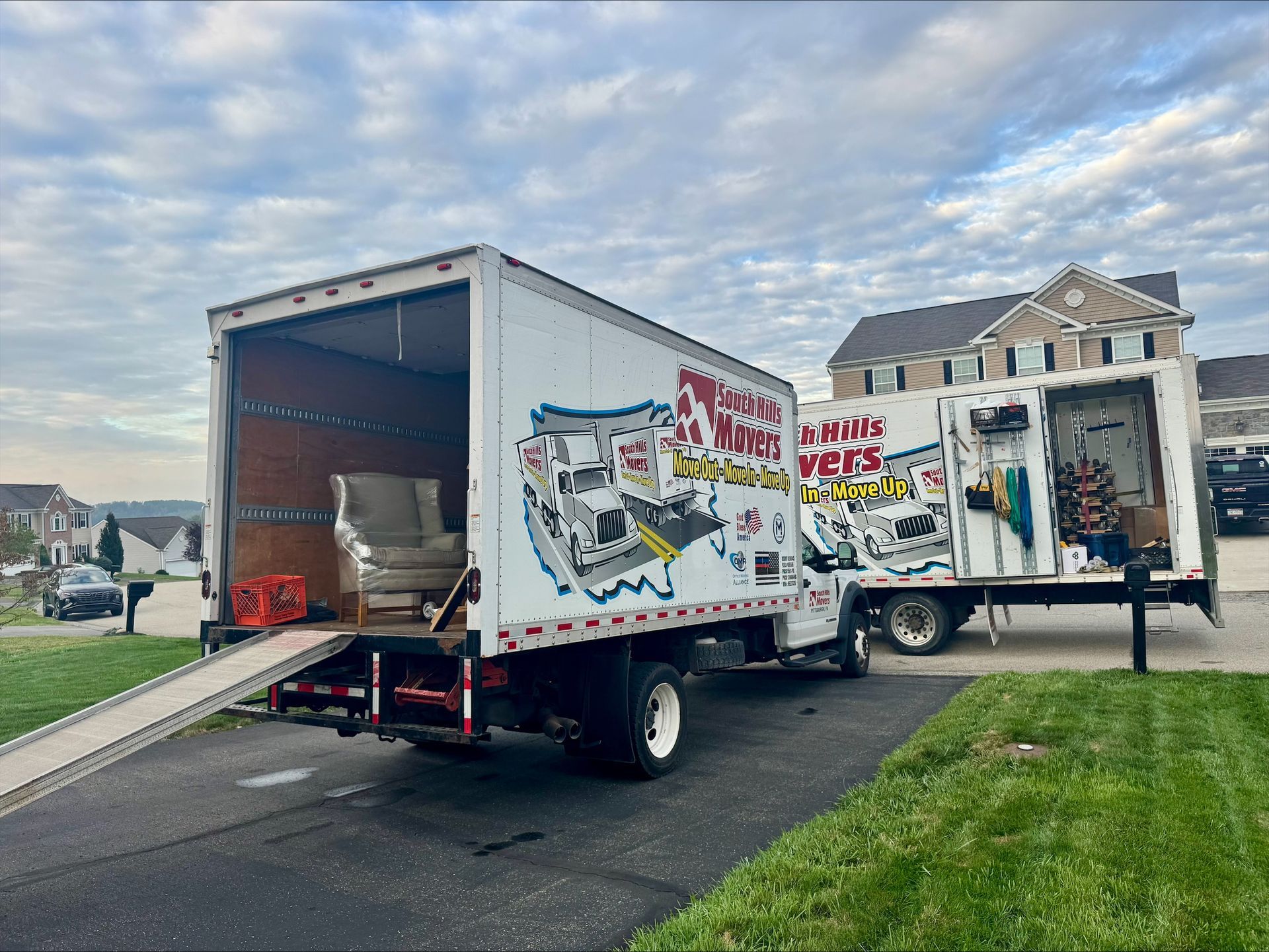 Moving truck parked in a driveway; ramp down, furniture inside, open doors, residential setting with cloudy sky.