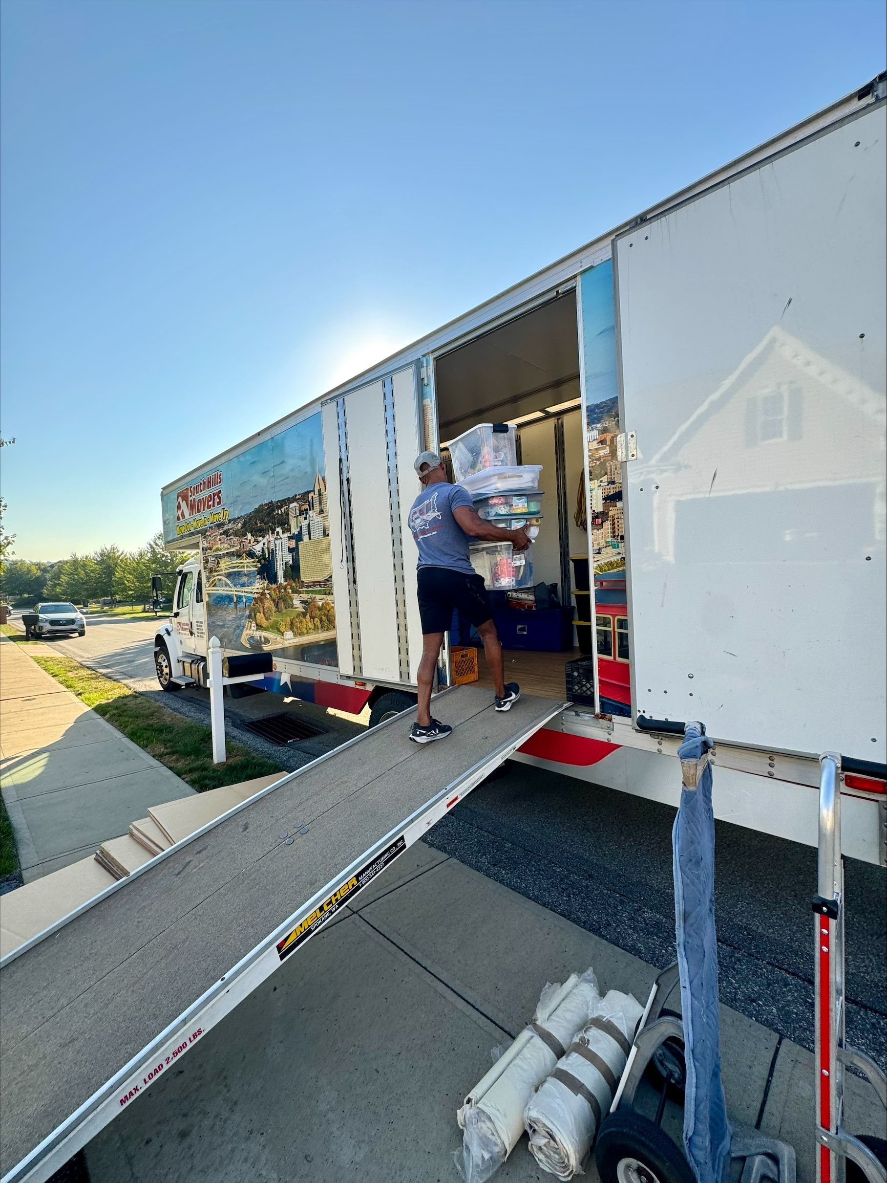 Man carrying boxes into a moving truck. A ramp is extended onto a driveway. Bright sunlight.